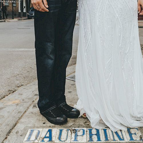 A couple stands on a street labeled "Dauphine" in a wedding dress and black pants, shoes, in an urban setting.