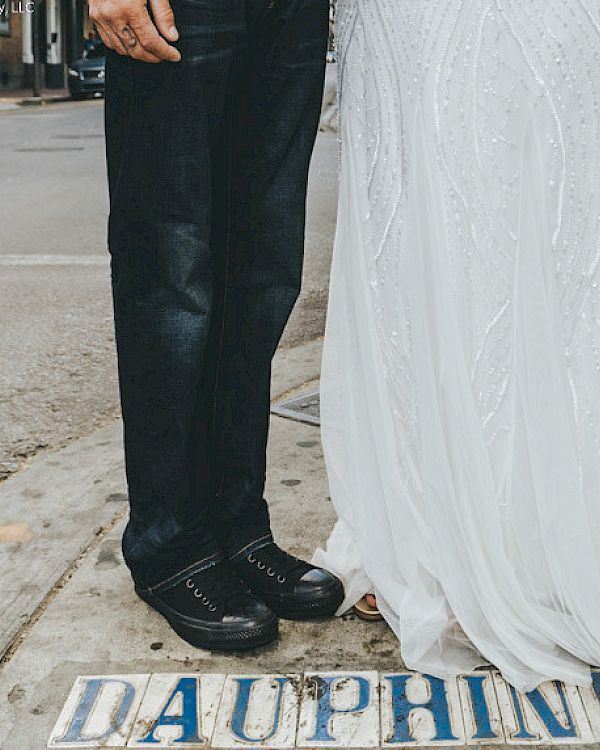 A couple stands on a street labeled "Dauphine" in a wedding dress and black pants, shoes, in an urban setting.
