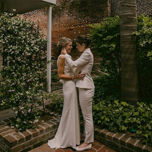 Two people in wedding attire lovingly gaze at each other in a lush garden setting with a brick wall and string lights in the background.