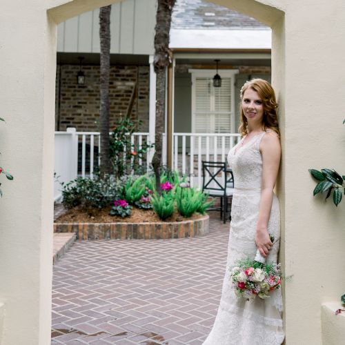 A woman in a white dress stands in an archway, holding a bouquet, with a garden backdrop.