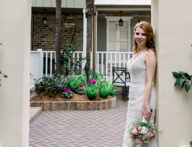 A woman in a white dress stands in an archway, holding a bouquet, with a garden backdrop.