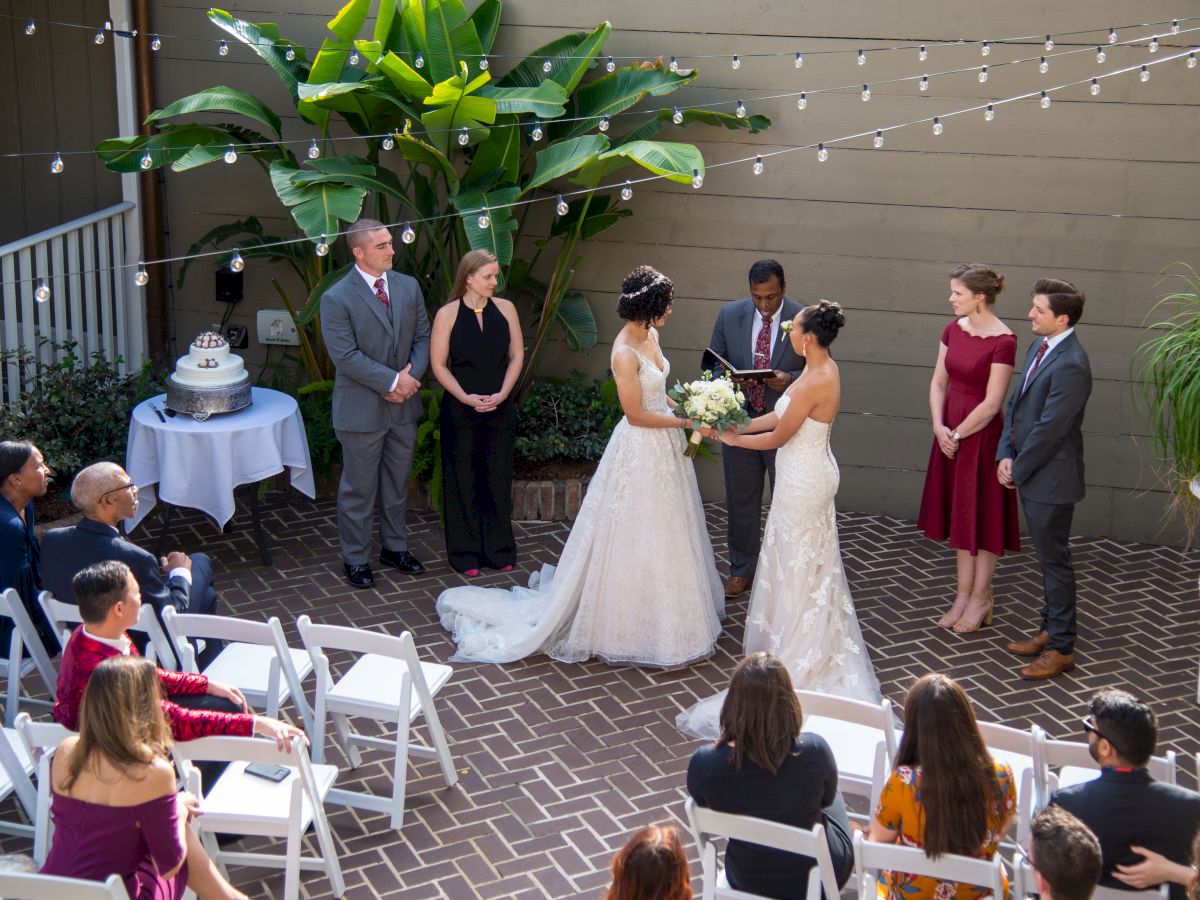 A wedding ceremony takes place outdoors with two brides in white gowns, surrounded by guests and a decorated setting.