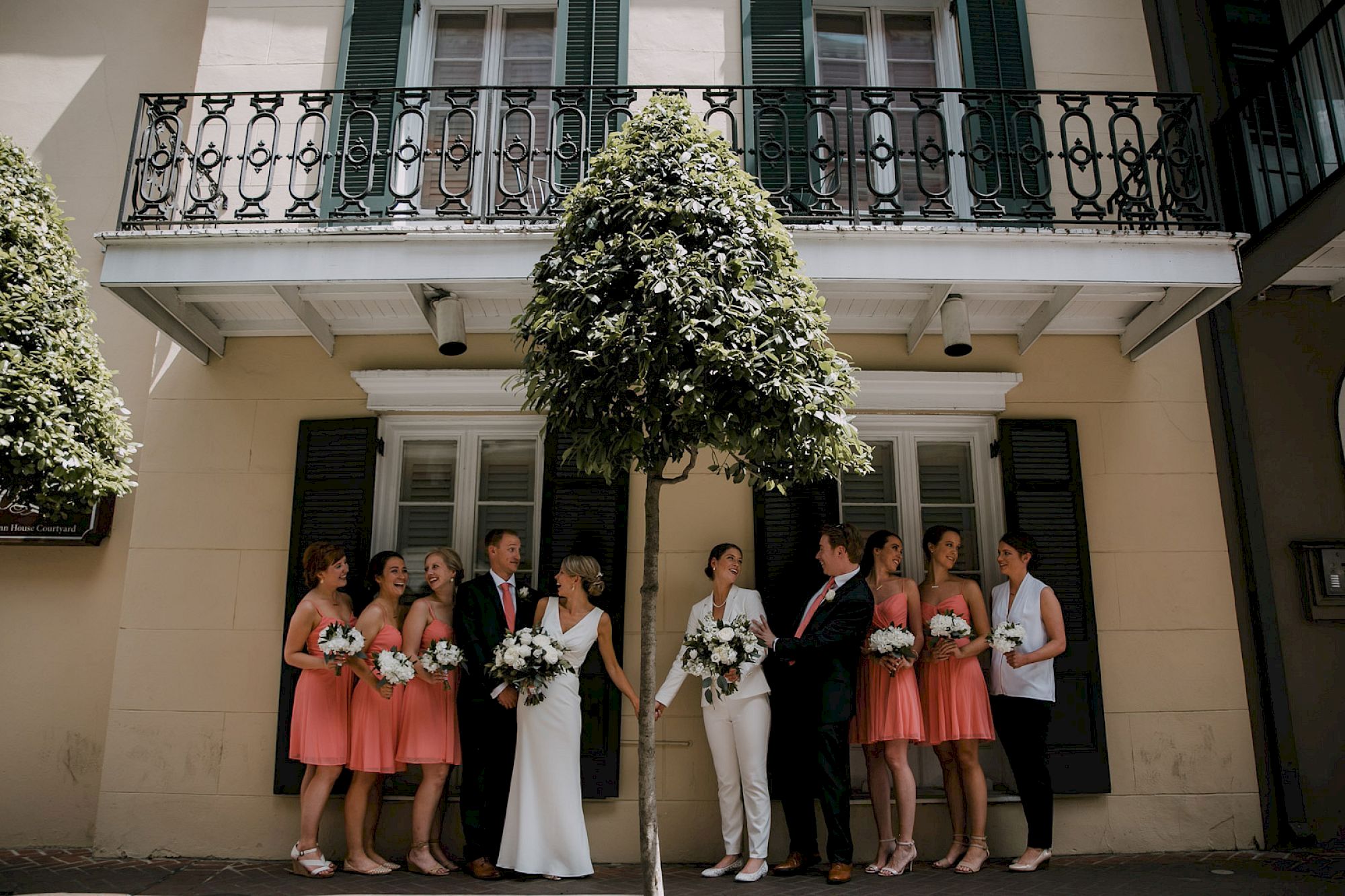 A wedding party stands outside a cream building with green shutters, bridesmaids in peach dresses, and the bride and groom under a balcony.