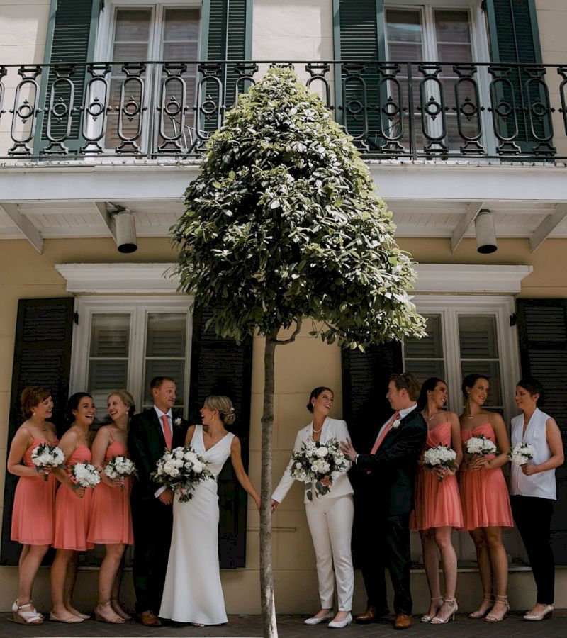 A wedding party stands in front of a building with a balcony and a tree.