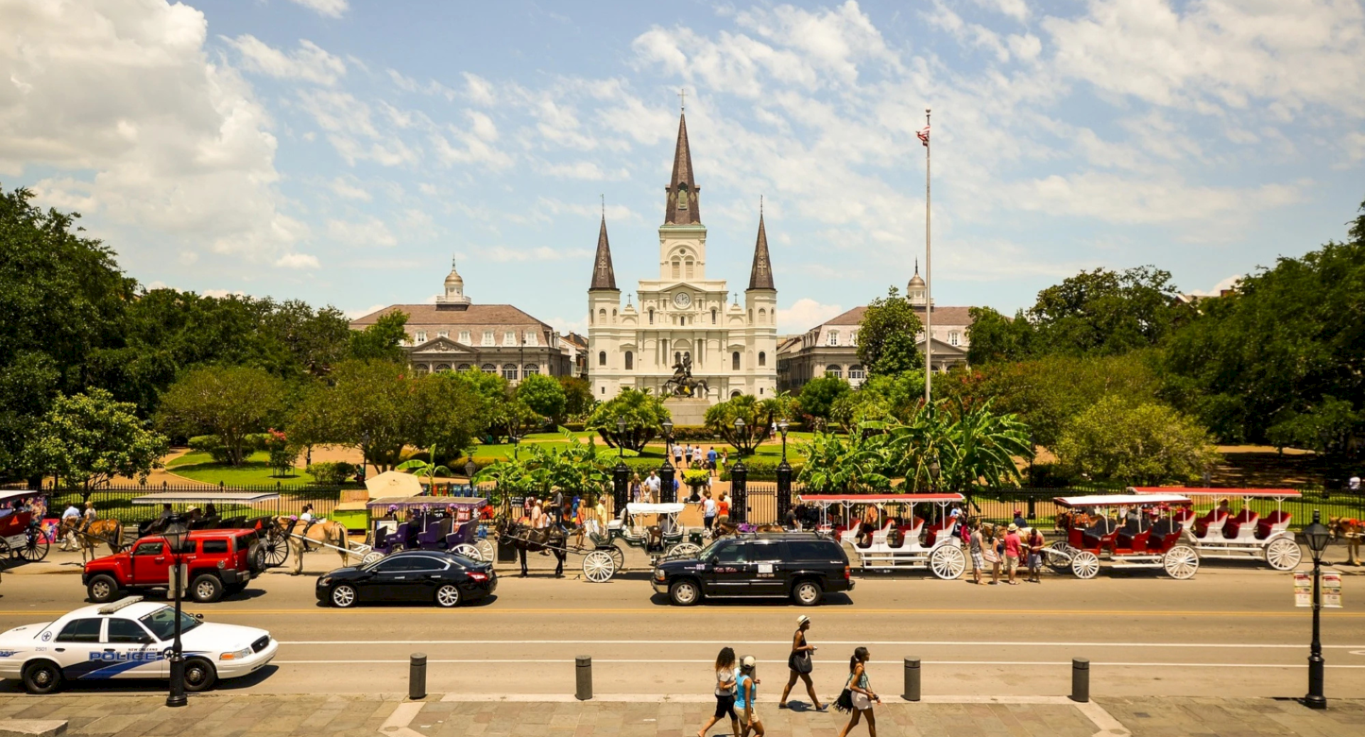 A grand white cathedral with tall spires sits behind a park, riding bikes and cars along a busy street in front of it.