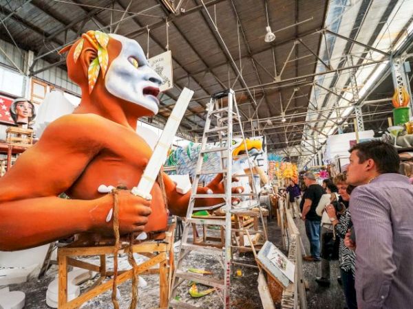 A worker with an orange costume and white mask is sculpting a large figure, holding a tool, in a busy workshop with onlookers.