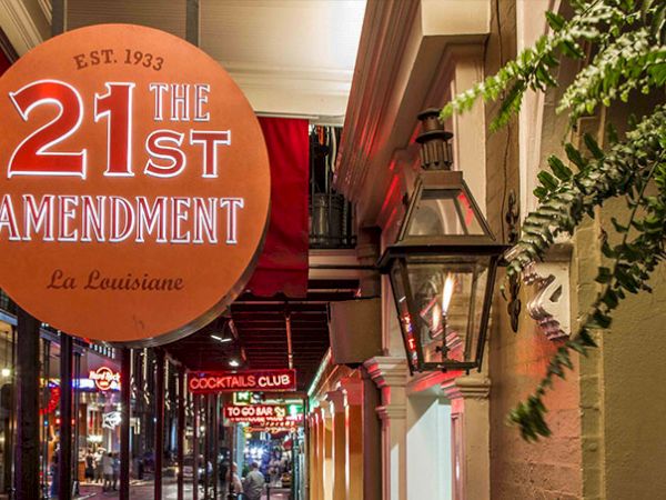 A street scene at night with a large circular sign reading &ldquo;21st The 1st Amendment&rdquo; and a lantern-lit storefront, Louisiana vibe, inset cocktail sign.