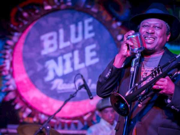 A smiling musician in a black hat and suit sings into a microphone on stage, holding a brass instrument, with a bright circular &ldquo;BLUE NILE&rdquo; sign in the background.