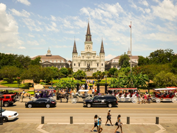 A sunny scene of a grand castle-like building with spires sits beyond a busy street; cars, cyclists, and pedestrians fill the foreground.