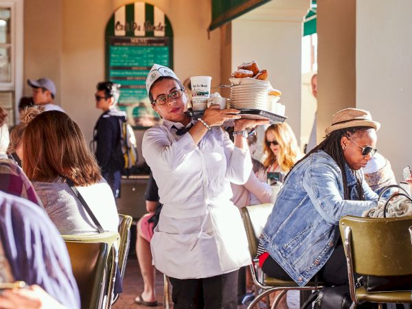 A waitress balanced two cakes on a tray while serving a busy outdoor cafe, with patrons at tables and warm hanging lights inside.