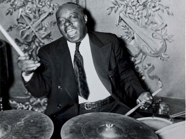 A black-and-white photo of a man playing a drum set, passionately playing sticks, wearing a suit and tie, indoors with ornate wall decor.