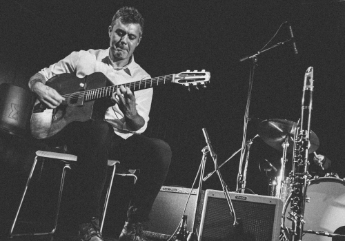A guitarist sits on a stool playing an acoustic guitar, with a drummer and saxes visible on a dim stage behind him.