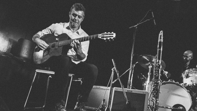A guitarist sits on a stool playing an acoustic guitar, with a drummer and saxes visible on a dim stage behind him.