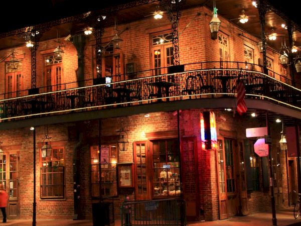 A glowing, two-story brick building at night with balcony lights, street lamps, and a corner storefront under warm lanterns.