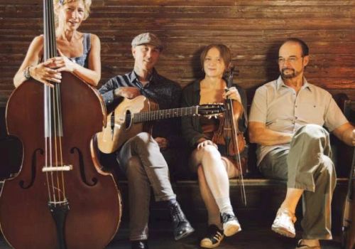 A group of four musicians sits on a stage with a cello, guitar, and two other string instruments, posing for a photo.