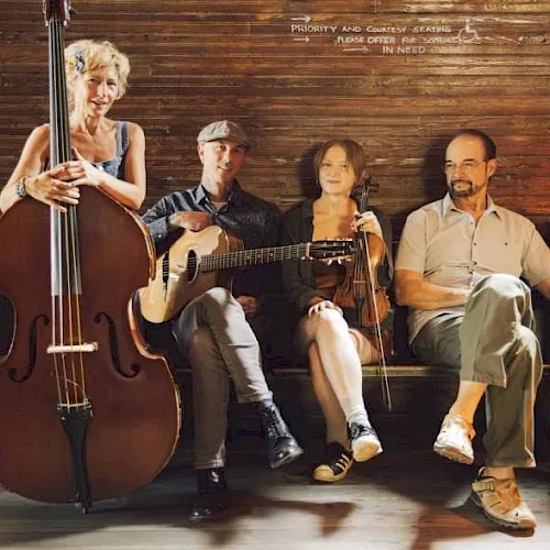 A group of four musicians sits on a stage with a cello, guitar, and two other string instruments, posing for a photo.
