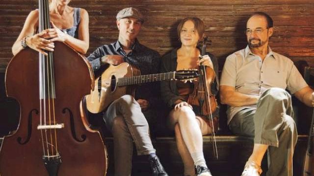 A group of four musicians sits on a stage with a cello, guitar, and two other string instruments, posing for a photo.