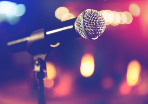 A silver studio microphone on a stand, with colorful blurred stage lights behind it.