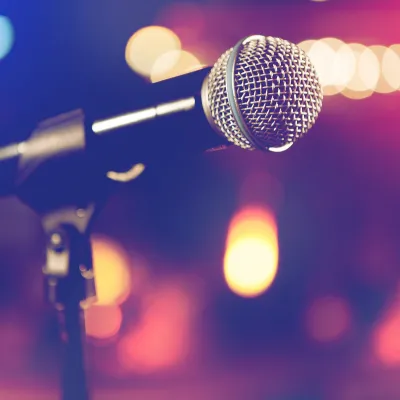 A silver studio microphone on a stand, with colorful blurred stage lights behind it.