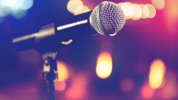 A silver studio microphone on a stand, with colorful blurred stage lights behind it.