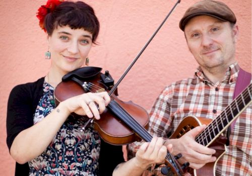 Two musicians pose against a pink wall, one plays violin with a bow while the other strums an acoustic guitar.