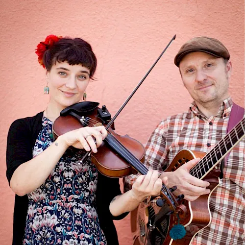 Two musicians pose against a pink wall, one plays violin with a bow while the other strums an acoustic guitar.