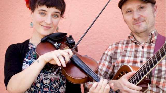Two musicians pose against a pink wall, one plays violin with a bow while the other strums an acoustic guitar.
