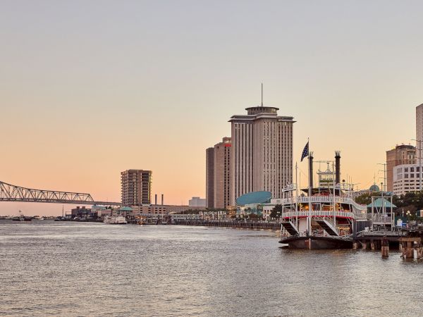A riverfront skyline with a tall tower building, an American flag on a docked riverboat, and bridges at sunset.