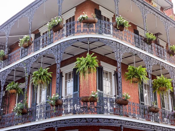 A red-brick building features ornate wrought-iron balconies packed with hanging flower pots and lush green plants.