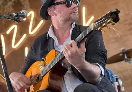 A musician in a fedora and sunglasses plays an orange electric guitar onstage, singing into a microphone amid warm lights.