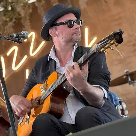 A musician in a fedora and sunglasses plays an orange electric guitar onstage, singing into a microphone amid warm lights.
