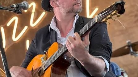 A musician in a fedora and sunglasses plays an orange electric guitar onstage, singing into a microphone amid warm lights.