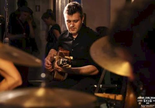 A man in a dim restaurant plays guitar while others watch, with warm stage lighting in the background.