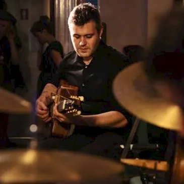 A man in a dim restaurant plays guitar while others watch, with warm stage lighting in the background.