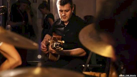 A man in a dim restaurant plays guitar while others watch, with warm stage lighting in the background.