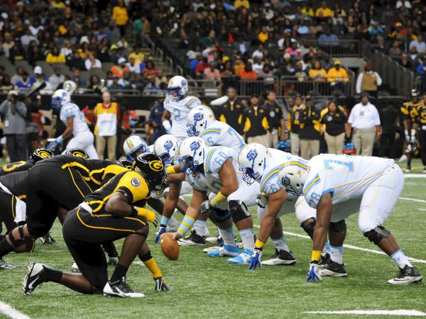 A football goal-line pileup as players in blue-and-white clash with defenders in black-and-yellow during a game.