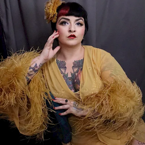 A made-up woman with dark hair and a gold feather boa, showing tattoos and striking a pose against a dark backdrop.