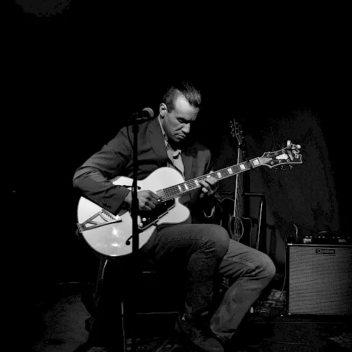 A black-and-white photo of a guitarist in a suit playing an acoustic-electric guitar while singing into a microphone on stage.