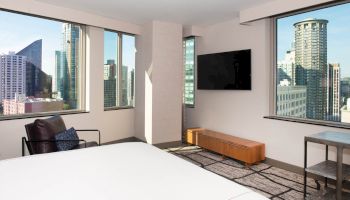 A modern bedroom with a city view features large windows, a bed, TV, chair, and a small table. Skyscrapers are visible outside.