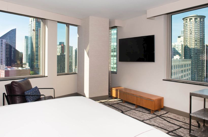 A modern bedroom with a city view features large windows, a bed, TV, chair, and a small table. Skyscrapers are visible outside.