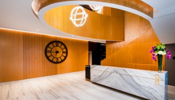 The image shows a modern reception area with wooden walls, a large clock, marble desk, and decorative lighting.