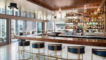 A modern bar with high stools, a fully stocked shelf of bottles, large windows, and a view of city buildings in the background.