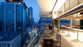 A modern cityscape featuring tall buildings and a stylish balcony with tables and chairs, overlooking a street at twilight.