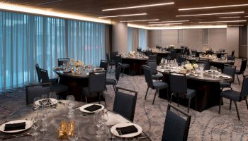 The image shows a modern, elegantly set dining area with round tables and chairs, prepared for an event with subtle lighting.