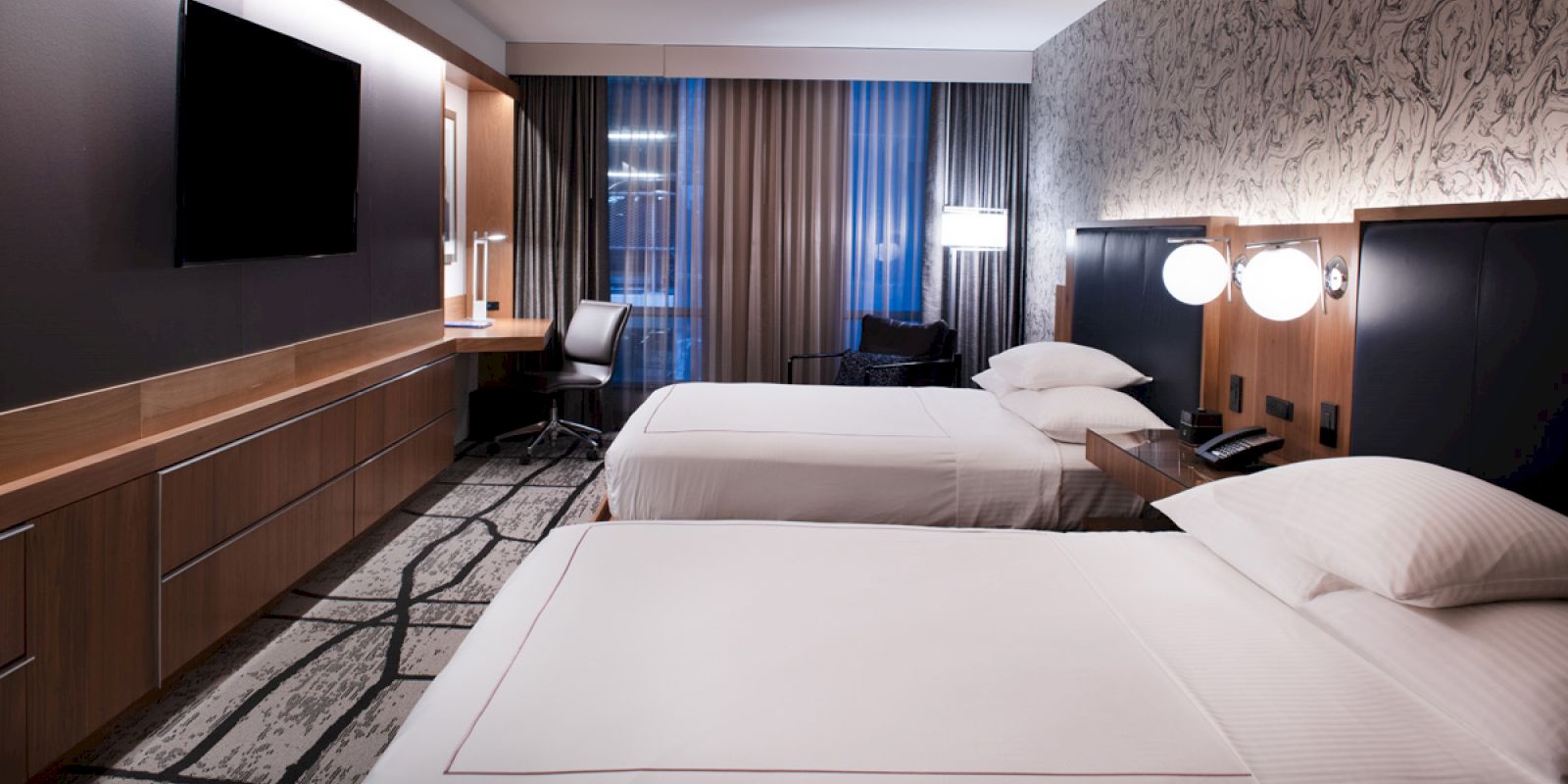 The image shows a modern hotel room with two beds, a large TV, a desk and chair, and stylish lighting and decor.
