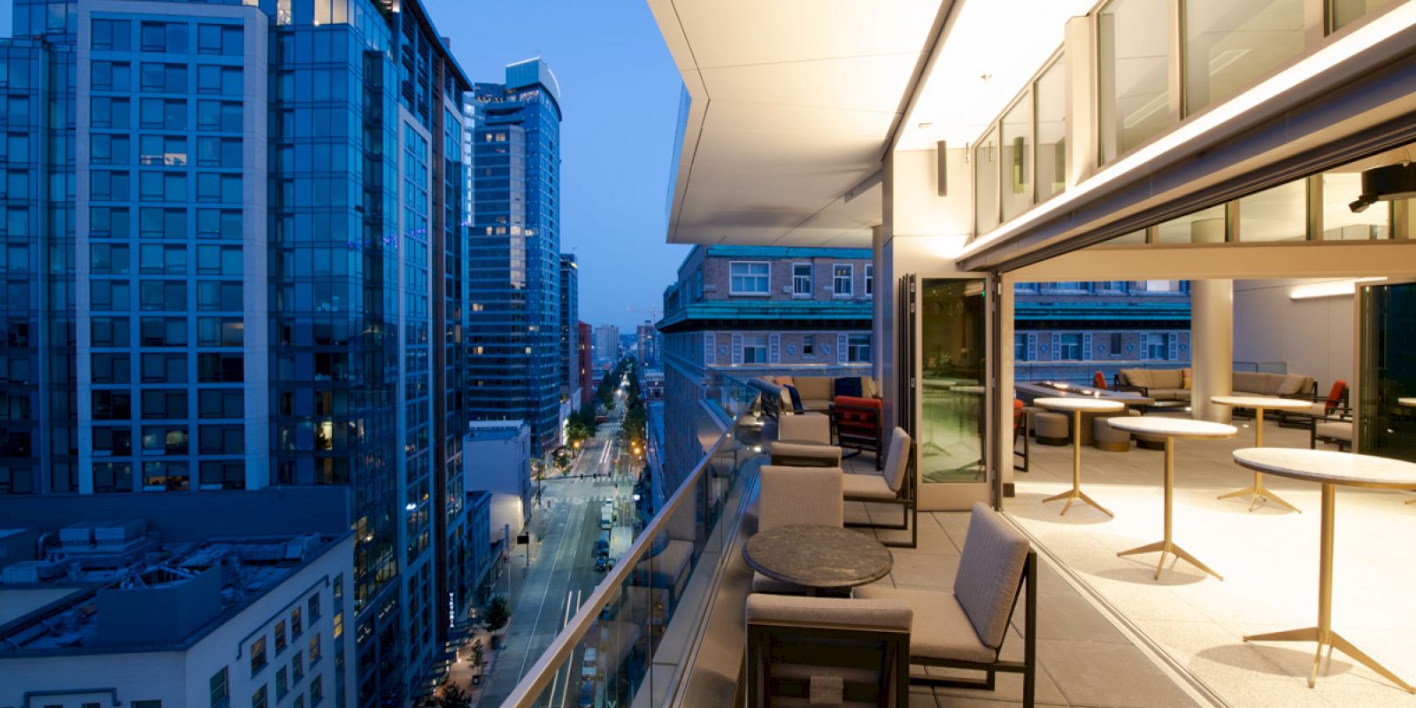 A modern balcony overlooks a city street at dusk, featuring outdoor seating and city skyscrapers in the background.