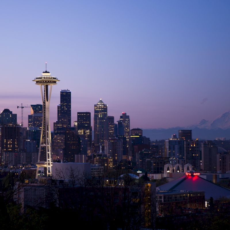 The image shows the Seattle skyline at dusk, featuring the Space Needle and Mount Rainier in the distance under a colorful sky.