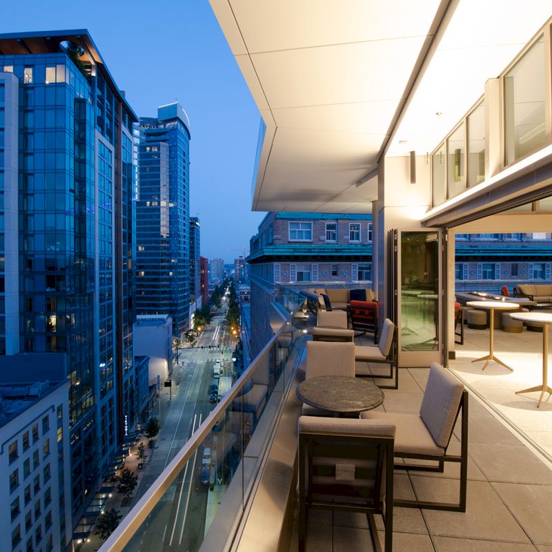 A modern cityscape at dusk, viewed from a balcony with tables and chairs, overlooking tall buildings and a lit street below.