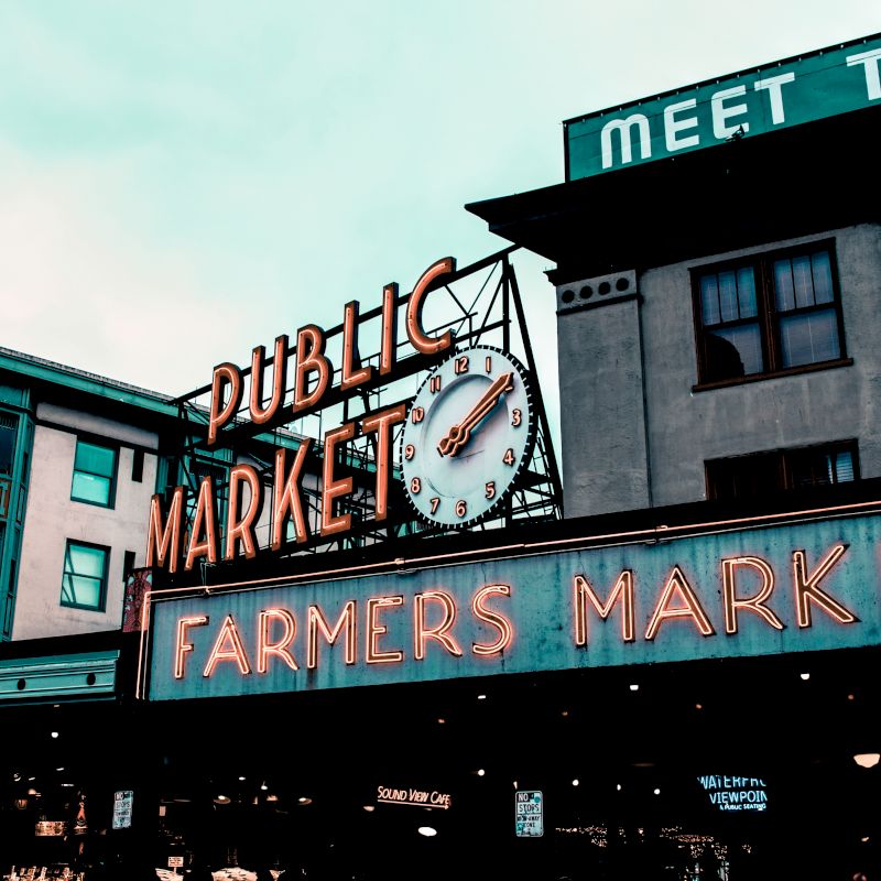 The image shows a sign for a public farmers market with a clock, set against buildings under a cloudy sky.