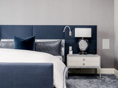 A modern bedroom features a blue upholstered headboard, white bedding, a stylish bedside table, and a contemporary lamp.